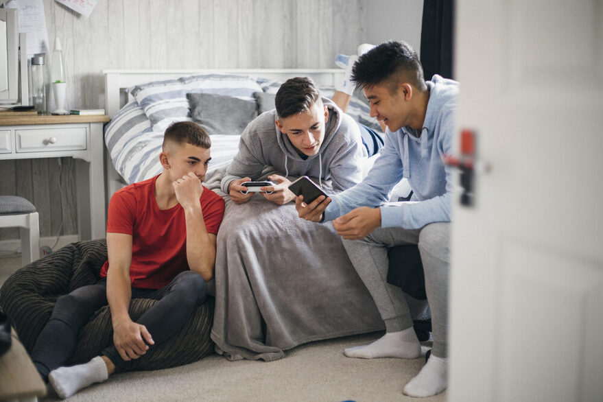 Drei männliche Jugendliche sitzen auf einem Bett und schauen in ein Smartphone.