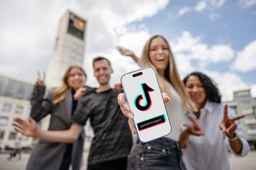 junge Menschen stehen vor dem Rathaus. In der Hand halten sie ein Smartphone mit dem TikTok Logo und dem Account-Namen der Stadt darauf. Der Account heißt @stadt.stuttgart