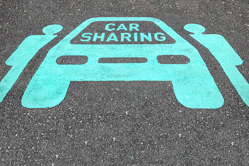 Ein hellblaues Piktogramm auf Asphalt zeigt zwei Männchen, die neben einem Auto stehen, auf dessen Windschutzscheibe "Carsharing" steht.