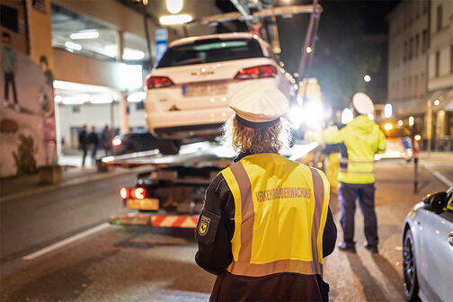 Eine Mitarbeiterin der Verkehrsüberwachung mit gelber Warnweste steht in der Stuttgarter Innenstadt abends vor einem weißen Pkw, der auf ein Abschleppfahrzeug gehoben wird.